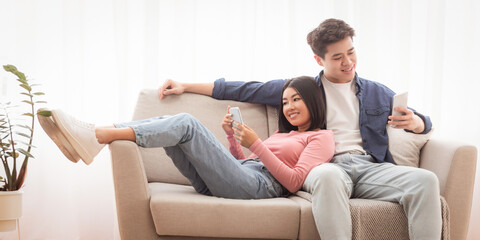 Korean Married Couple With Smartphones Browsing Internet And Using Apps Relaxing Sitting On Couch In Living Room At Home. Gadget Lifestyle, Mobile App Concept. Free Space For Text