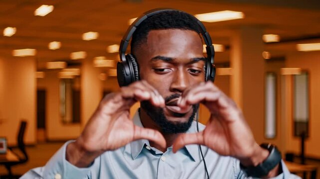 Confident young man listening to music with headphones, making heart gesture with hands in modern
