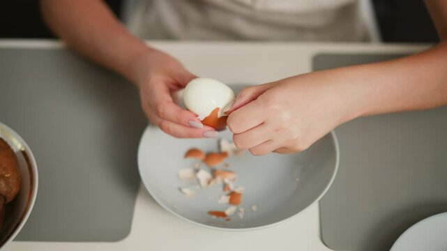 Overhead view of fair skinned person focused on peeling shell off boiled egg over bowl on table with scattered shell fragments, on placemat beside potatoes and empty dish