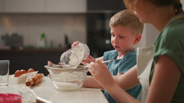 boy holds transparent jar of flour, pouring it into sieve held by woman while looking up and speaking to her, surrounded by baking ingredients and utensils on kitchen counter
