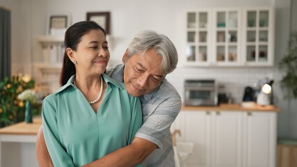 Fototapeta premium Happy senior couple with casual cloth spending time together and hugging at modern kitchen. Grandmother and grandfather holding hand together and looking at camera. Healthy relationship. Myrmidon.
