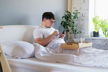 Young man waking up and having breakfast in bed and using smartphone