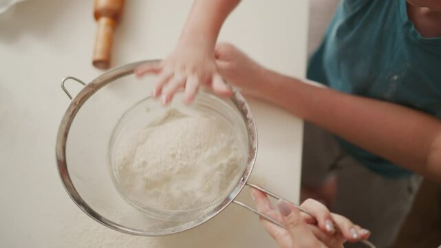 overhead view of child pressing flour through sieve into glass bowl while adult supports from side on white countertop with baking tools like rolling pin visible