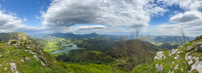 Breathtaking panoramic view embracing lush green valley, serene lake and majestic mountains under cloudy sky.
