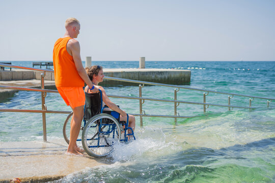 Woman with disability, entering the sea water with the help of a lifeguard assistant across an accessible wheelchair beach ramp, rear view. - Powered by Adobe