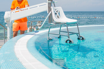 Man handling a pool chair lift, lifting and lowering from and into the water to provide access to the swimming pool for people with a physical disability.