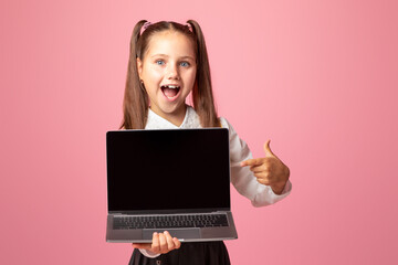 Naklejka premium Wow, look there. Happy little girl with open mouth shows at laptop with blank screen, isolated on pink background, copy space