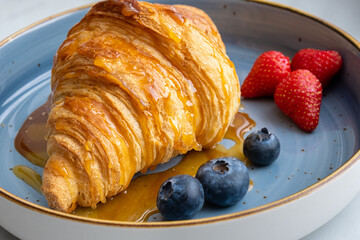 Close-up of a golden croissant covered in thick sweet maple syrup, with fresh blueberries and strawberries nearby. Tasty high-calorie breakfast or a delightful dessert to start the day