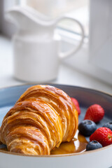 Golden crispy croissant drizzled with maple syrup, served on a blue plate with fresh strawberries and blueberries. French breakfast with cappuccino, milk jug in background. Close-up shot.