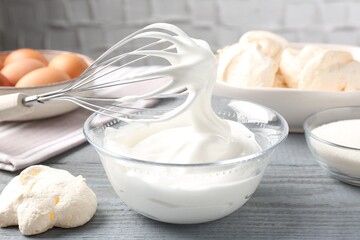 Whisk with whipped cream and other ingredients on gray wooden table, closeup. Cooking meringue...