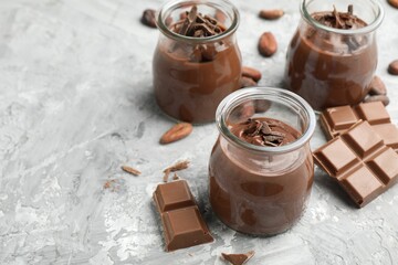 Tasty pudding in glass jars, pieces of chocolate and cocoa beans on light grey table, closeup. Space for text