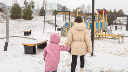 Young mother and a little girl in warm outerwear walk in the park in the children's playground in...