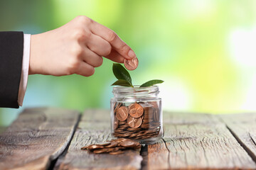 Investment concept. Woman putting coin into glass jar at wooden table against blurred green...