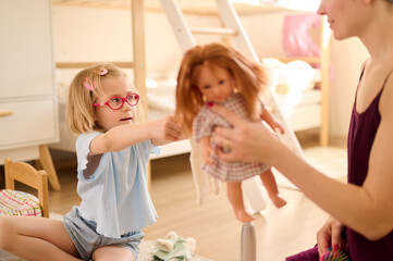 child playing with doll bonding moment in cozy room with natural light. little girl in glasses actively engaged, building creativity and imagination. childhood, family interaction, playtime