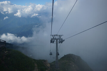  A cable car glides above green mountains shrouded in mist, creating a serene atmosphere. The landscape is breathtaking, showcasing clouds and distant peaks.