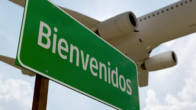 4k Bienvenidos Green Road Sign and Airplane Flying Low Overhead with Landing Gear Down.
