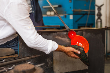 An artisan in a Murano workshop manipulates a glowing orange piece of molten glass with tools,...