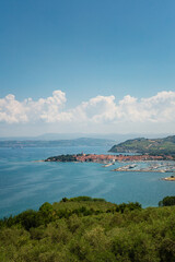 A scenic panoramic view of the coastal town of Izola in Slovenian Istria, featuring the harbor, red-tiled roofs, and the blue Adriatic Sea under a partly cloudy sky.