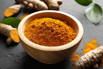 Aromatic turmeric powder in bowl, fresh roots and leaves on black table, closeup