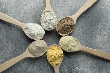 Different types of flour in spoons on grey table, flat lay