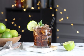 Tasty Cuba Libre cocktail in glass, limes and tongs on white marble table, bokeh effect