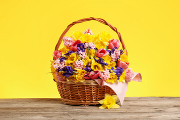 Wicker basket with beautiful flowers on wooden table against yellow background