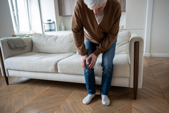 Unrecognizable disabled pensioner trying stand up from sofa with swollen arthritic knees. Rheumatoid joint damage, stiffness, movement restriction, chronic inflammation, progressive deformity