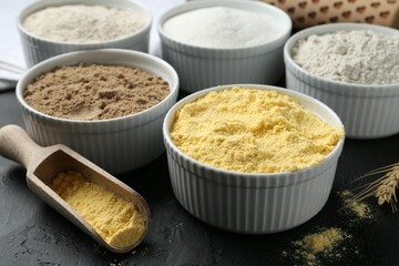 Different types of flour in bowls and scoop on black textured table, closeup