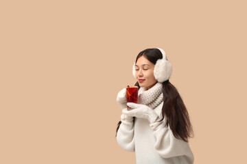 Beautiful young happy Asian woman in winter clothes with cup of hot mulled wine on beige background