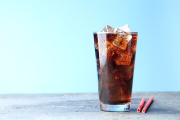 Refreshing cola with ice cubes in glass and straws on grey textured table against light blue gradient background, closeup. Space for text