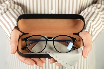 Woman holding case with stylish glasses and cleaning cloth on light background, closeup