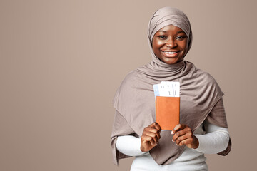 Cheerful black islamic woman in headscarf posing with passport and tickets over light studio...
