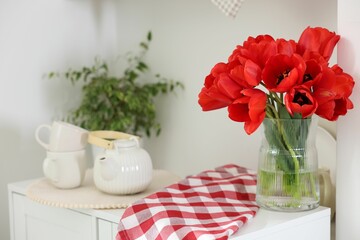 Beautiful red tulips in vase, cups and teapot on cabinet indoors