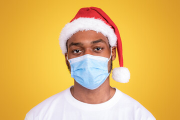 Coronavirus Christmas. Sad African American Man Wearing Covid-19 Face Mask And Santa Hat Celebrating Winter Holidays And New Year Alone During Pandemic Posing On Yellow Studio Background
