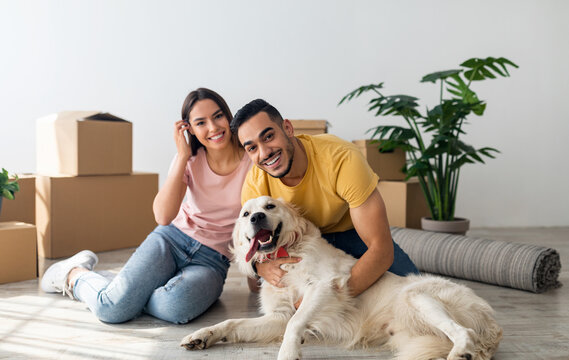 Full length portrait of happy young diverse couple with their dog posing on floor of new home on moving day. Millennial homeowners with cute pet sitting among cardboard boxes, relocating to new house