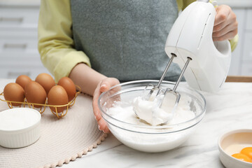 Woman whisking cream with hand mixer at white marble table indoors, closeup