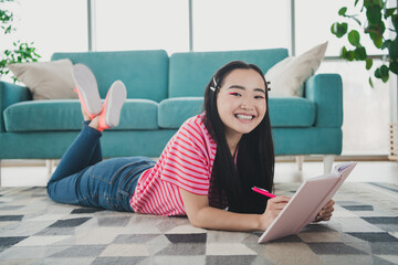 Cheerful young woman relaxing indoors on a sunny day, lying on a soft rug with a notebook,...