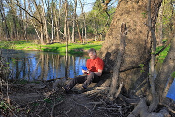 Middle-aged man in a red shirt finds a peaceful place by a stream to read