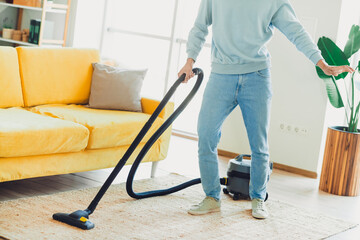 Fototapeta premium Young man vacuuming living room carpets at home while enjoying casual indoor time