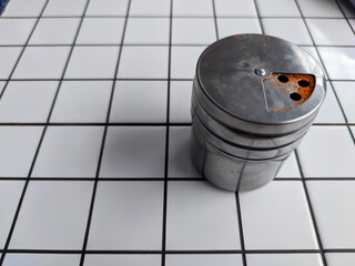A silver chili powder dispenser sits on a black-and-white checkered tile surface. Reddish residue near the dispensing holes indicates its contents.