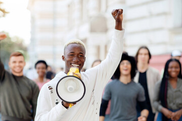 Emotional black guy activist with megaphone on the street, chanting slogans and raising fist up,...