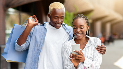 Shopping online and in mall. Cheerful african american couple with many colored packages look at smartphone and make new purchase, free space