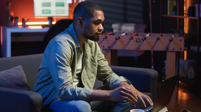 Young black guy working on his freelance tasks in the living room, adjusting his job schedule and enjoying work from his cozy home. Person navigating on web with laptop, multitasking. Camera B.