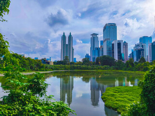 Jakarta City skyline reflected in calm water, framed by lush greenery under a partly cloudy sky, blending urban architecture with nature.