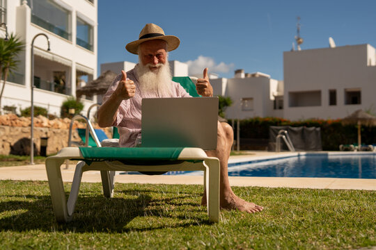 Smiling retired man making a video call on his laptop while sitting by the pool at a Spanish resort, enjoying relaxed retirement life and digital communication on vacation - Powered by Adobe