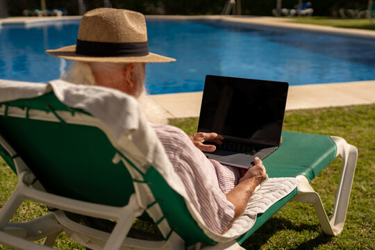 Elderly freelancer working remotely on laptop while relaxing on sun lounger near outdoor swimming pool at resort, enjoying peaceful retirement and digital nomad lifestyle