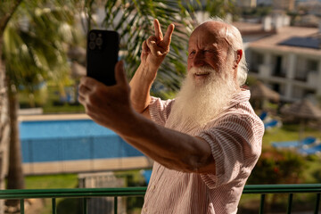 Happy senior man with long white beard taking selfie with smartphone on balcony, enjoying sunny vacation in tropical resort