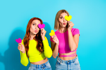 Two playful young women holding colorful heart-shaped cards against a vibrant turquoise background