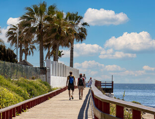 two people walking along ocean on wooden boardwalk with palm trees and sky