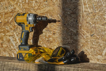 Cordless drill, yellow safety goggles, and gloves rest on a wood surface with a particle board...